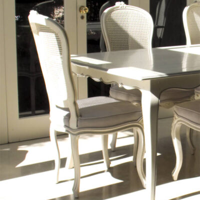 French provincial white painted chair upholstered with cane back next to with white dining table by window with sunlight